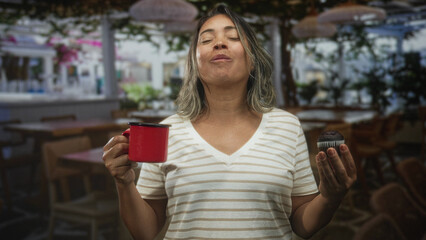 Woman holding red cup in right hand and chocolate muffin in left hand, smiling in a cafe building;...