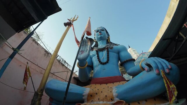A large, vibrant blue statue of Lord Shiva in a sitting meditative pose on a tiger skin statue  with a Trishul and sacred flags inside a temple complex.