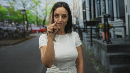 Fototapeta premium Woman pointing index finger to camera on street, white tshirt with visible hand and forearm gesture near face conveying emphasis; confidence.