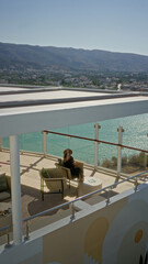 Fototapeta premium Woman sitting holding sunhat by glass railing overlooking turquoise sea from cruise terrace building with wicker chairs and small table in sunlight; serenity.