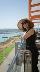 Fototapeta premium Woman smiling leaning on glass railing on cruise deck overlooking harbor and coastline; serenity vacation.