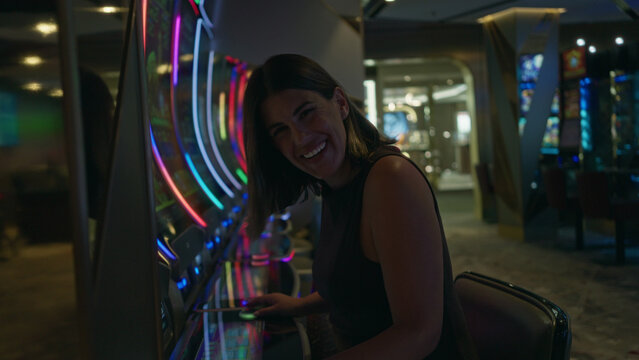 Woman brunette sitting with hand on slot machine control panel, smiling broadly and looking up toward the screen in a casino building; luck excitement.