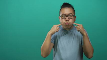 Fototapeta premium Young chinese man with glasses puffs his cheeks and points both index fingers at his cheeks in a studio with a teal green backdrop; playful mischief.