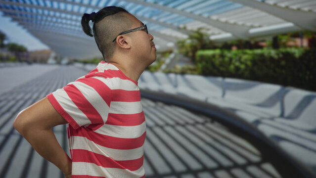 Young chinese man with glasses and manbun wearing red striped shirt leaning forward and holding lower back for backache on street under louvered canopy; discomfort.
