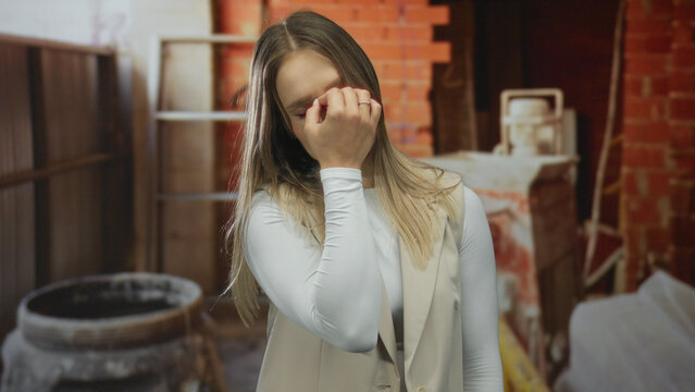 Woman indoors at construction site appearing worried with blonde hair wearing white top and beige vest surrounded by industrial equipment and unfinished walls displaying concern.