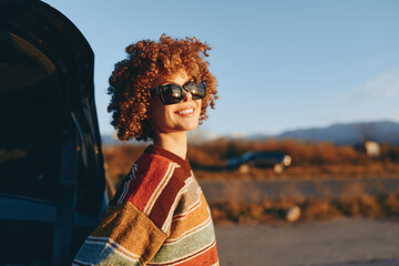 Smiling woman with curly hair wearing sunglasses and rainbow sweater stands near car outdoors in warm sunlight, enjoying lifestyle moment with positive mood and casual style. © SHOTPRIME STUDIO