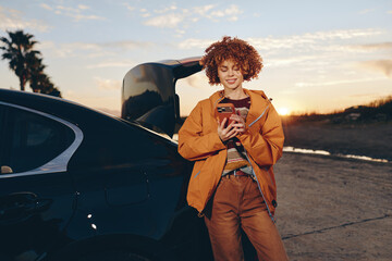 Young woman with curly hair wearing rainbow sweater and orange jacket stands by car trunk outdoors...