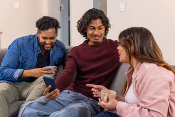 Naklejka premium Asian friends in denim and sweaters laughing and pointing at smartphone on gray sofa with mugs