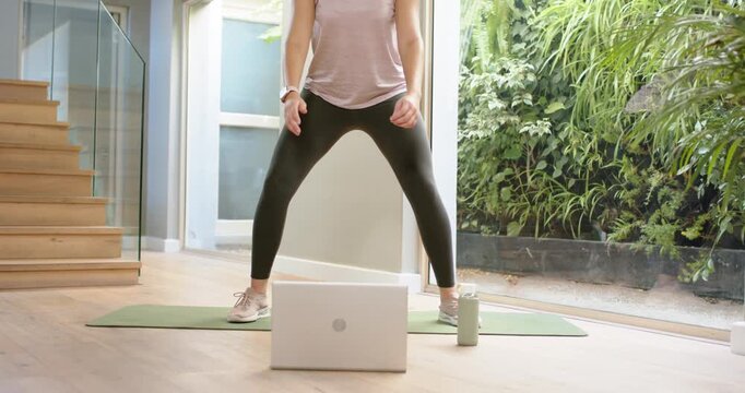 African American woman following laptop workout, lowering and rising into deep plie squat at home
