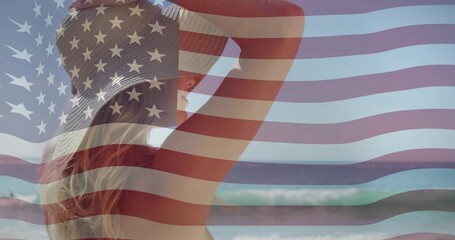 Naklejka premium Standing woman watching waves at beach, wearing straw hat, swimsuit, US flag overlay, copy space