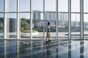 Fototapeta premium Man standing in front of glass curtain wall while looking at his smartphone