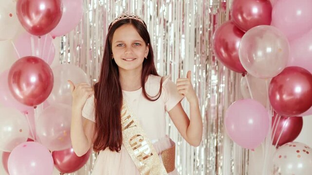 A young girl stands in a party area surrounded by balloons and shiny decorations. She smiles and waves while wearing a birthday sash that shows her special day