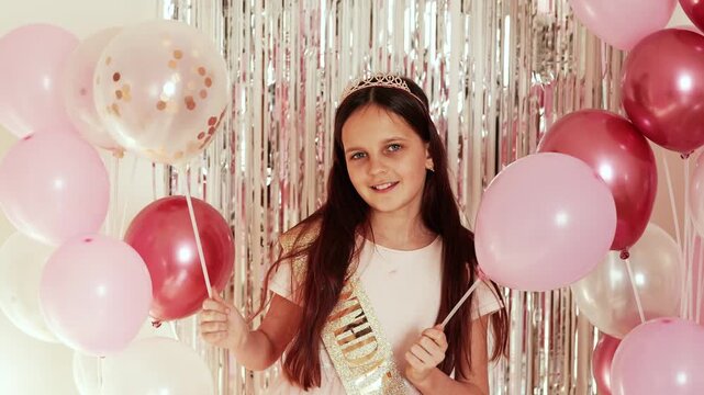 A girl celebrates her birthday. She holds pink and clear balloons in front of a shiny backdrop. She smiles while wearing a party sash and a crown