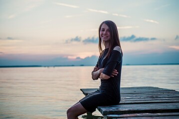 Fototapeta premium Young woman portrait sitting on wooden pier by lake at sunset