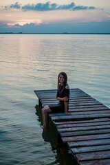 Fototapeta premium Smiling young woman sitting on wooden pier over calm lake at sunset