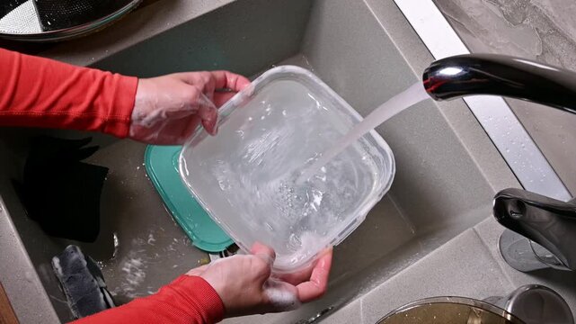 Caucasian woman washing plastic container under running water in kitchen sink, hands soapy, with dish soap and sponge visible in the background