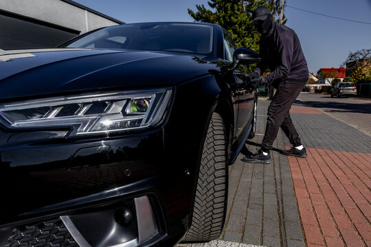 A masked man tries to steal a luxury car from the parking lot of an apartment, a car thief dressed in black with a hat and a balaclava covering his eyes,