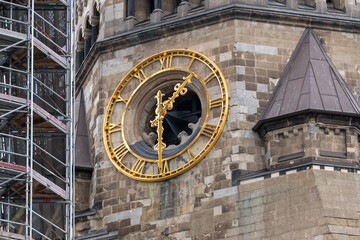 Obraz premium Stone tower features a large golden clock face with Roman numerals. The clock shows gold hands indicating the time. Scaffolding stands to the left of the tower. Roman numerals mark each hour clearly