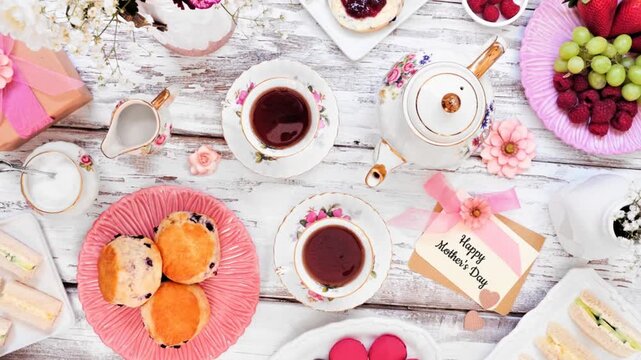 Mothers Day tea table scene over a white wood background. Slow rotating zoom motion. Greeting card, gift, flowers, vintage tea set, finger sandwiches, desserts and fruit.