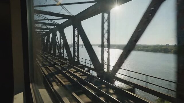 Perspective view from a moving train window crossing a massive steel truss bridge over a wide river with bright sunlight reflecting on the water surface.
