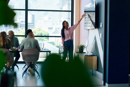 Young businesswoman in smart casual pointing at screen while presenting to team in bright office