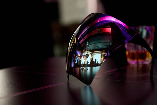Futuristic reflective visor mask with purple neon light on dark table. Mirror surface reflects party scene with champagne glasses and people. Creative cyberpunk nightlife and masquerade concept.