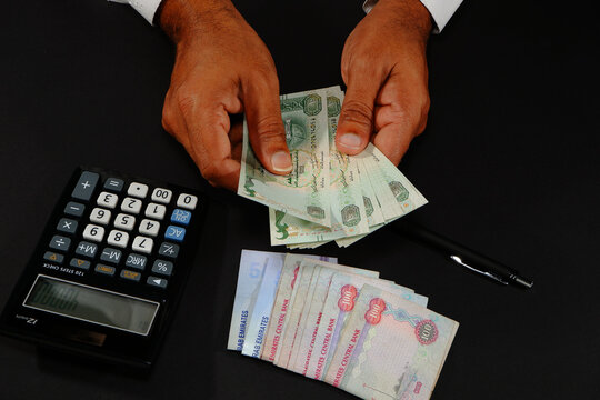 Close-up high angle view of hands counting stacks of UAE Dirham banknotes next to a black electronic calculator displaying numbers and a pen on black surface