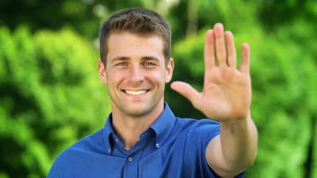 A smiling man in a blue shirt holding up his hand in a stopping motion outdoors with greenery in the background, possibly for a gesture or warning sign