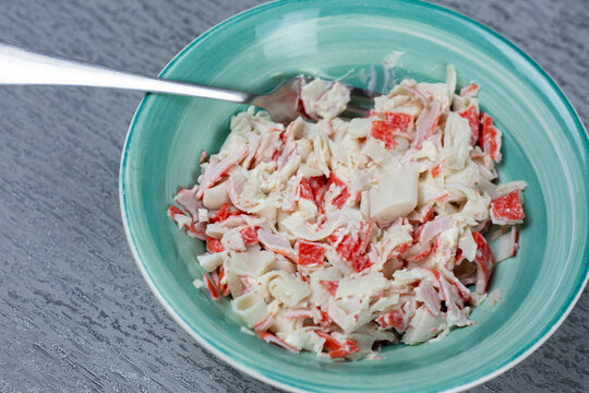 Bowl filled with crab salad featuring imitation crab meat, mayonnaise, and diced vegetables, placed on a textured gray surface with a fork positioned on the edge of the bowl