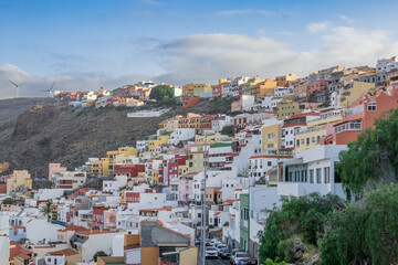 Fototapeta premium San Sebastián de la Gomera: pintorescas casas de colores escalonadas sobre la ladera de la montaña. Paisaje urbano canario con molinos eólicos, cielo suave y arquitectura típica de la isla.
