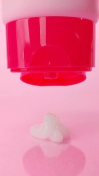 Squeezing or dispensing white beauty serum from a bright pink cosmetic bottle or tube. Isolated on matching pink background, skincare product presentation.