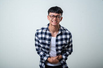Fototapeta premium Young Asian man wearing round glasses, plaid shirt, and white tank top bends forward while clutching his stomach and screaming in pain against a light gray background