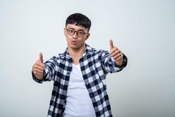 Fototapeta premium Young Asian man with round glasses wearing a plaid shirt and white tank top showing thumbs up while smiling broadly in front of a light gray background