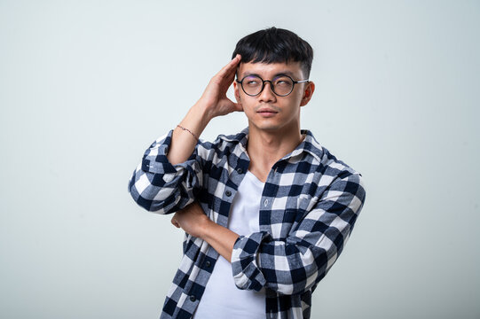 Young Asian man wearing round glasses, plaid shirt, and white tank top holds his head with one hand while grimacing in pain against a light gray background