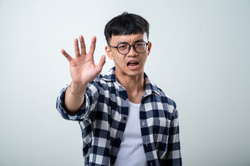 Naklejka premium Young Asian man wearing round glasses, plaid shirt, and white tank top extends his hand forward in a stop gesture while standing against a light gray background