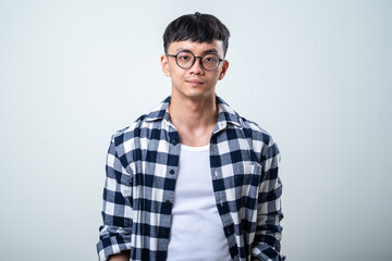 Young Asian man wearing round glasses, a plaid shirt, and white tank top stands against a light gray background, looking at the camera with a calm, casual expression © Daniel