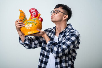 Young Asian man in round glasses and a plaid shirt holds a colorful rooster-shaped decorative...