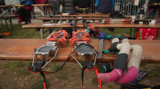 Orange crampons and ice axes rest on a wooden bench, prepared for an ice climbing competition. In the background, a crowd of spectators and participants gathers, creating a focused sporting atmosphere