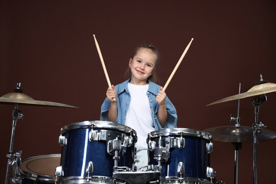 Little girl with drumsticks and drum kit on brown background