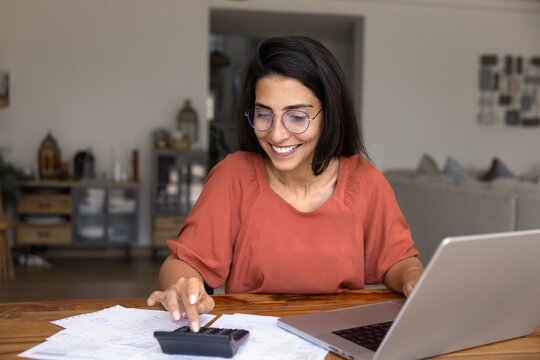 Smiling young woman in glasses calculate finances at home, using calculator and laptop seated at desk with documents and receipts, feeling satisfied with budget results, sufficient incomes and savings