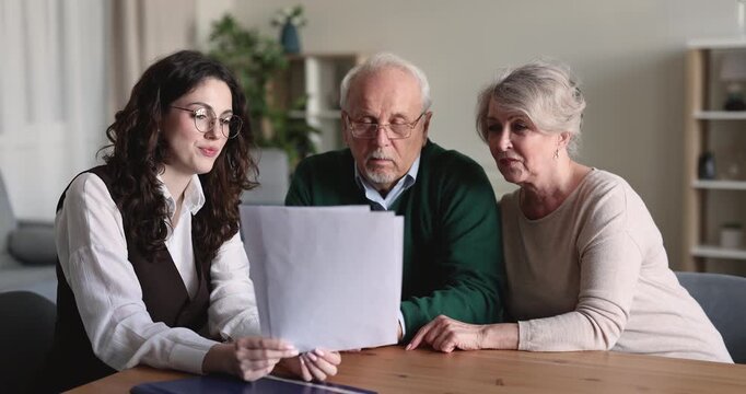 Young adult female advisor explaining retirement plan, discussing medical insurance coverage policy, reviewing loan contract or mortgage financial documents with senior couple during home consultation