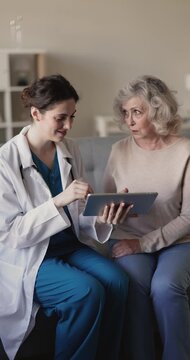 Vertical view female doctor holding digital tablet, explains e-health application use, share medical information or check-up results to senior woman during home consultation. Healthcare, telemedicine