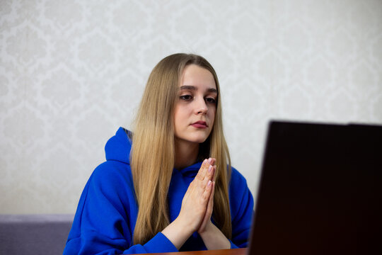 Young blonde woman prays with hands clasped while watching online church service on laptop. Digital worship and spiritual connection through internet broadcast at home. Faith goes virtual