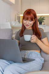 Young woman at home holds coffee cup checks email on laptop works freelance on tasks and replies....