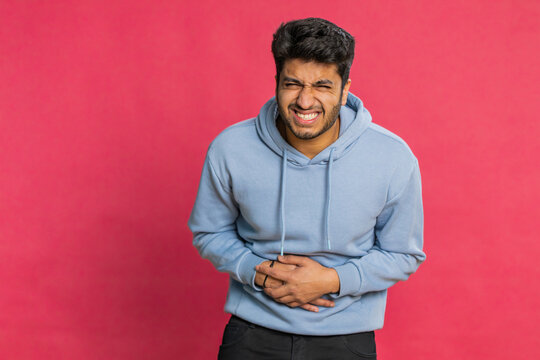 Indian man feeling sudden strong abdominal stomach ache, gastritis problem. Arabian guy having symptom poisoning diarrhea indigestion peptic ulcer pancreatitis isolated on red background indoors