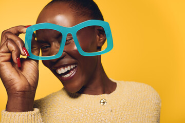 Happy dark skinned African woman smiling broadly holds oversized blue glasses near face. Yellow...