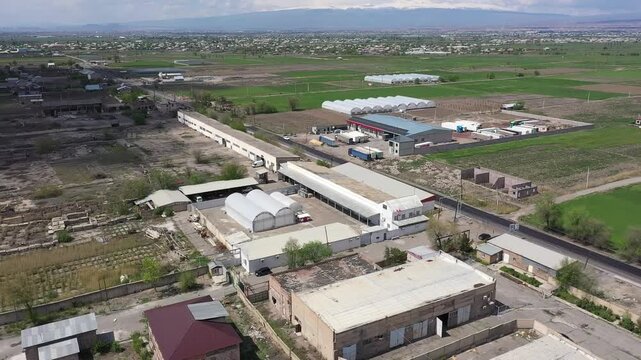 Aerial drone footage showing a large agricultural complex with multiple industrial buildings and a massive greenhouse situated in a rural landscape with green fields and a distant town.