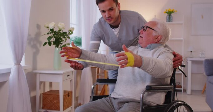 Senior man in wheelchair doing exercise using rubber band with support from nurse. Young caregiver physiotherapist helping elderly man with in rehabilitation in retirement home. 4k video.