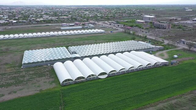 Aerial drone footage showing a large agricultural complex with multiple industrial buildings and a massive greenhouse situated in a rural landscape with green fields and a distant town.