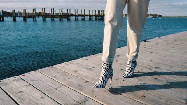 Lady in white overall and zebra high-heeled boots walks by the pier. Happy smiling woman in orange glasses holds her big hat.